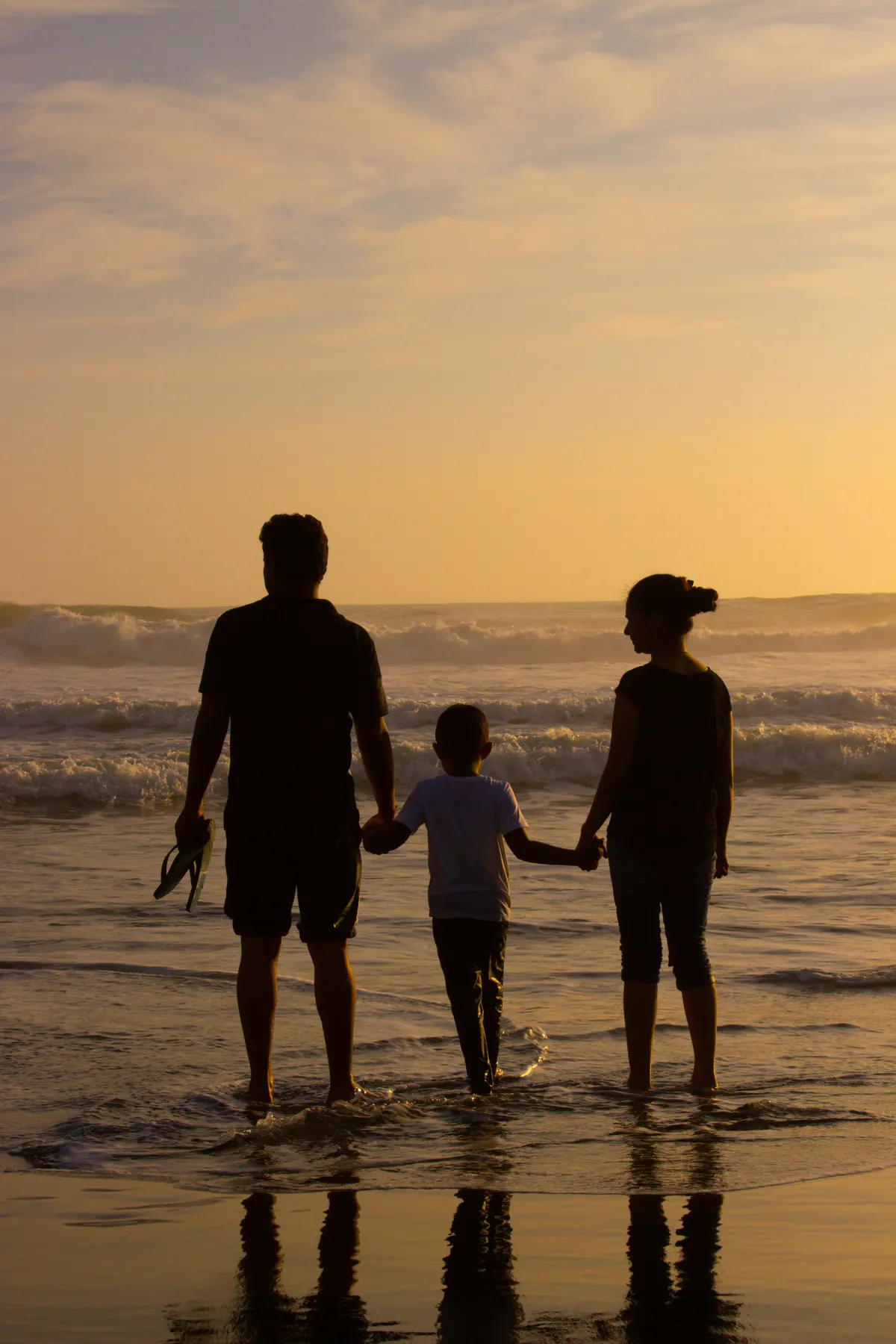 An adult holding hands with two children at the water's edge, seen in silhouette