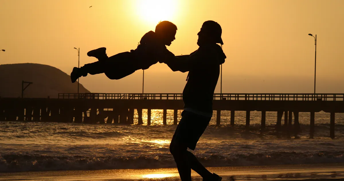 An adult swinging a child by the arms on a pier at sunset, seen in silhouette