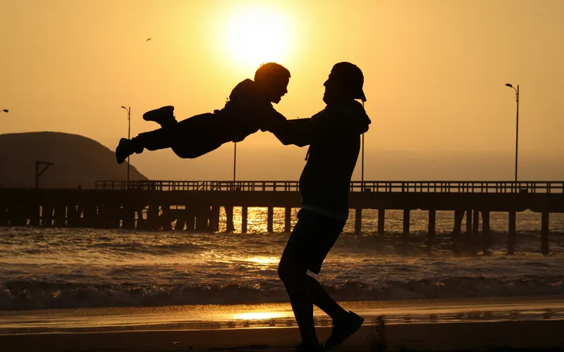 An adult swinging a child by the arms on a pier at sunset, seen in silhouette