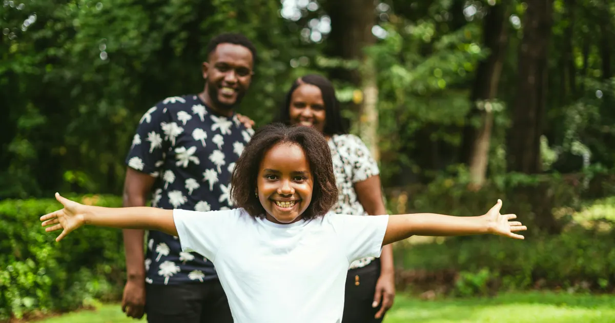 A child standing with arms outstretched on a lawn, two parents behind