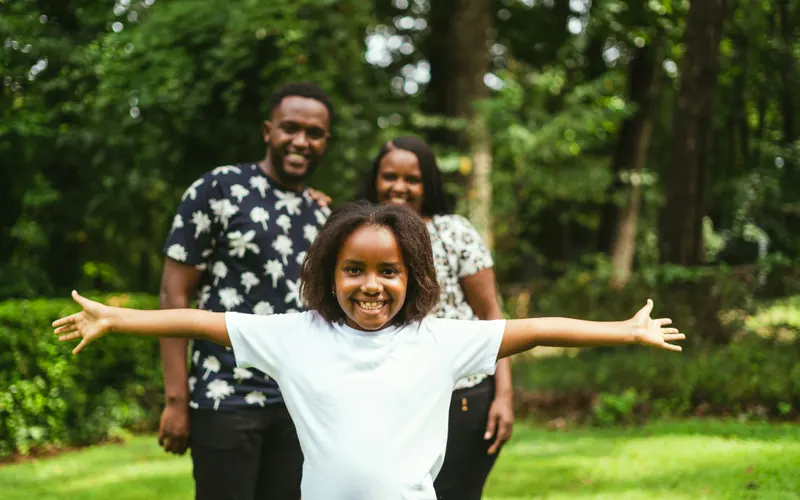 A child standing with arms outstretched on a lawn, two parents behind