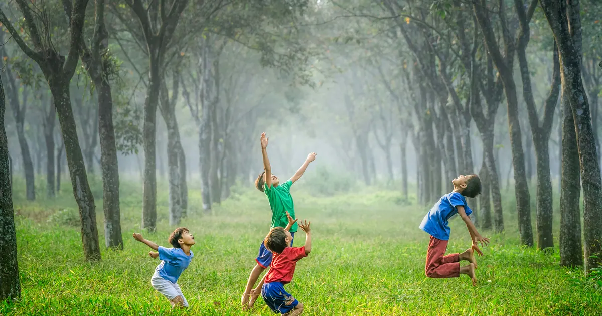 Three children playing with a ball on grass between two rows of tall trees