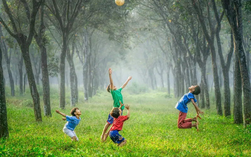Three children playing with a ball on grass between two rows of tall trees