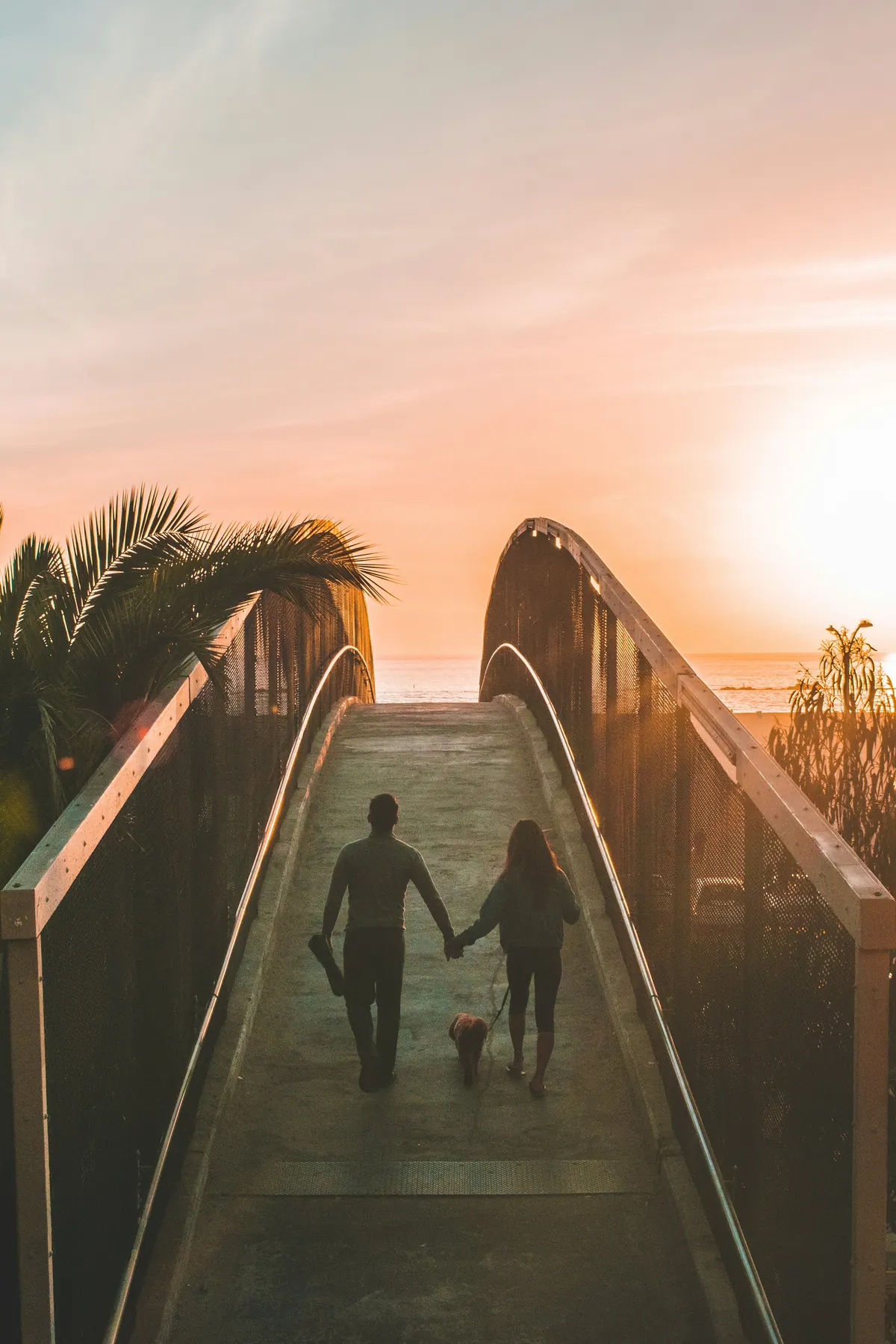 A couple walking a dog across a wooden footbridge towards the sea at sunset
