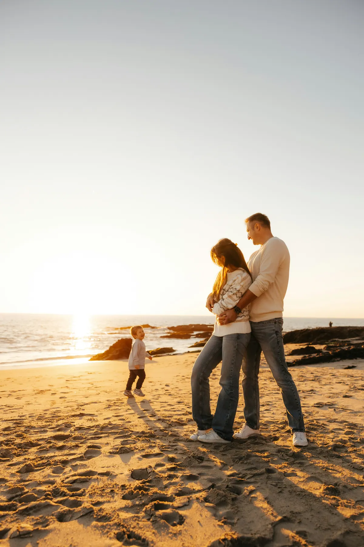 A couple embracing on a beach at sunset while a small child plays nearby