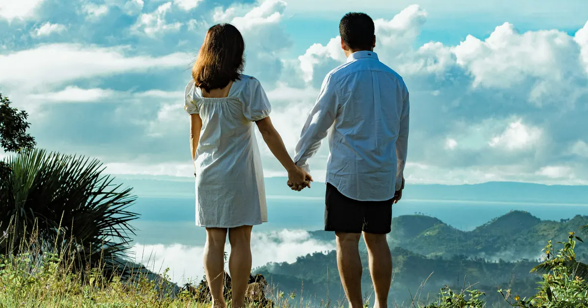 A couple standing hand in hand on a hillside, looking out towards distant mountains