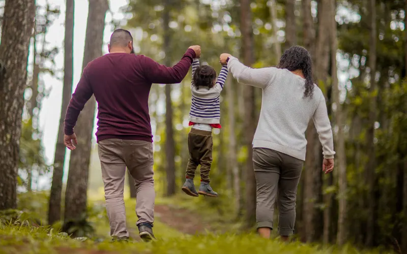 Two parents walking a woodland path swinging a small child between them