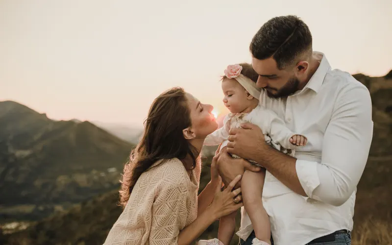 Two parents holding a baby between them on a hillside at sunset, mountains behind