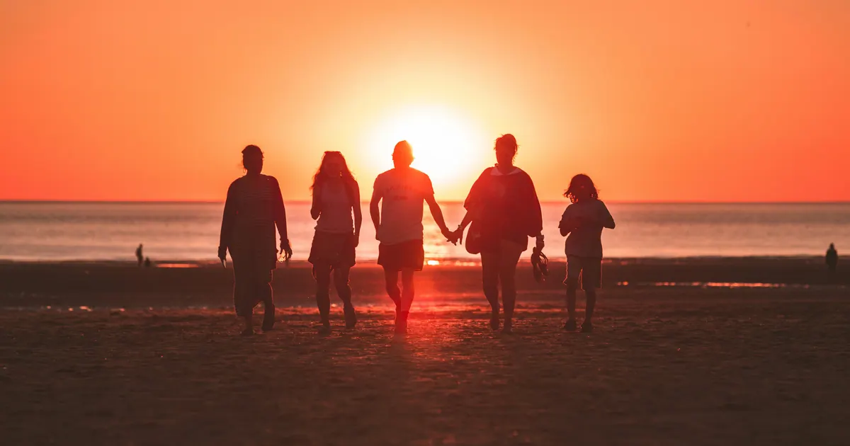 A family of five walking along a shoreline at sunset, seen in silhouette