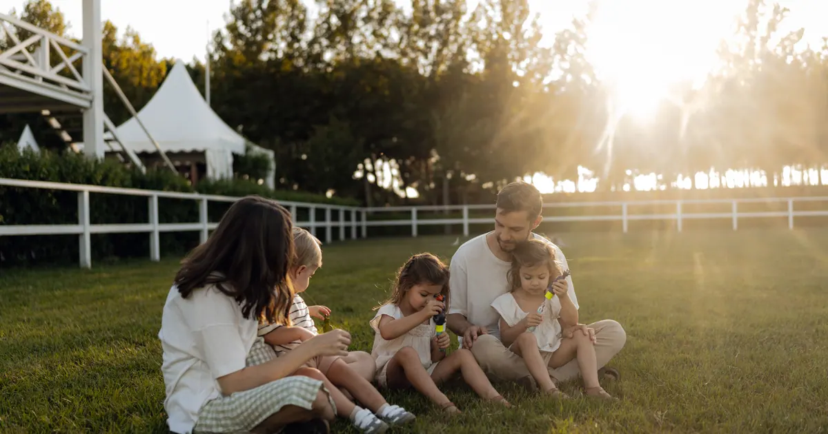 A family of four sitting on grass beside a white fence in evening light