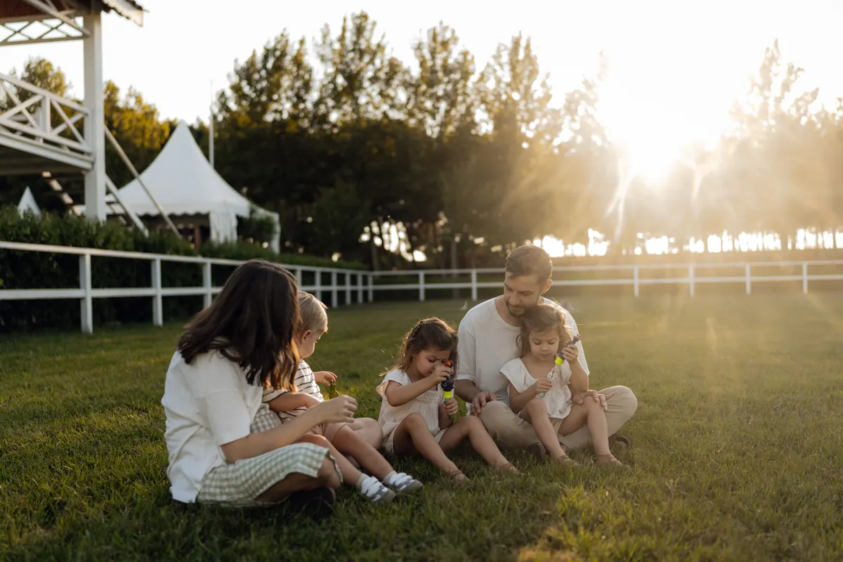 A family of four sitting on grass beside a white fence in evening light