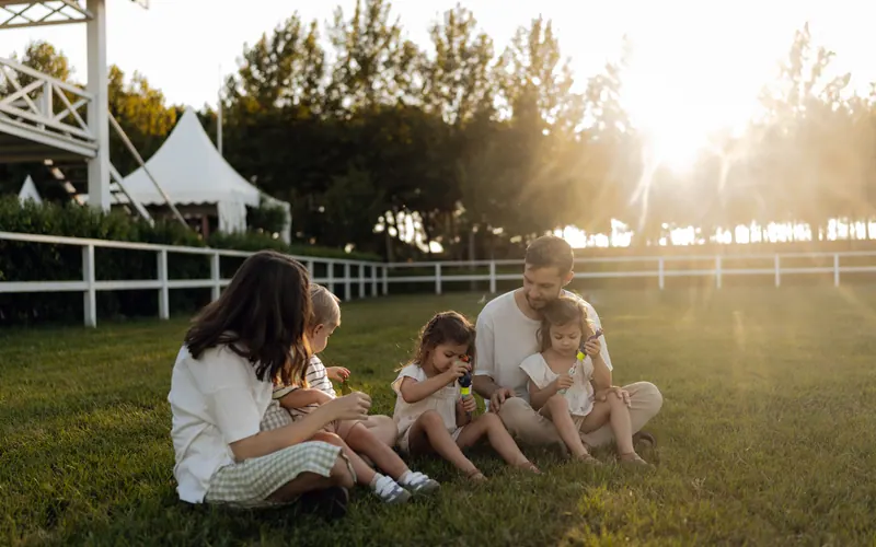A family of four sitting on grass beside a white fence in evening light