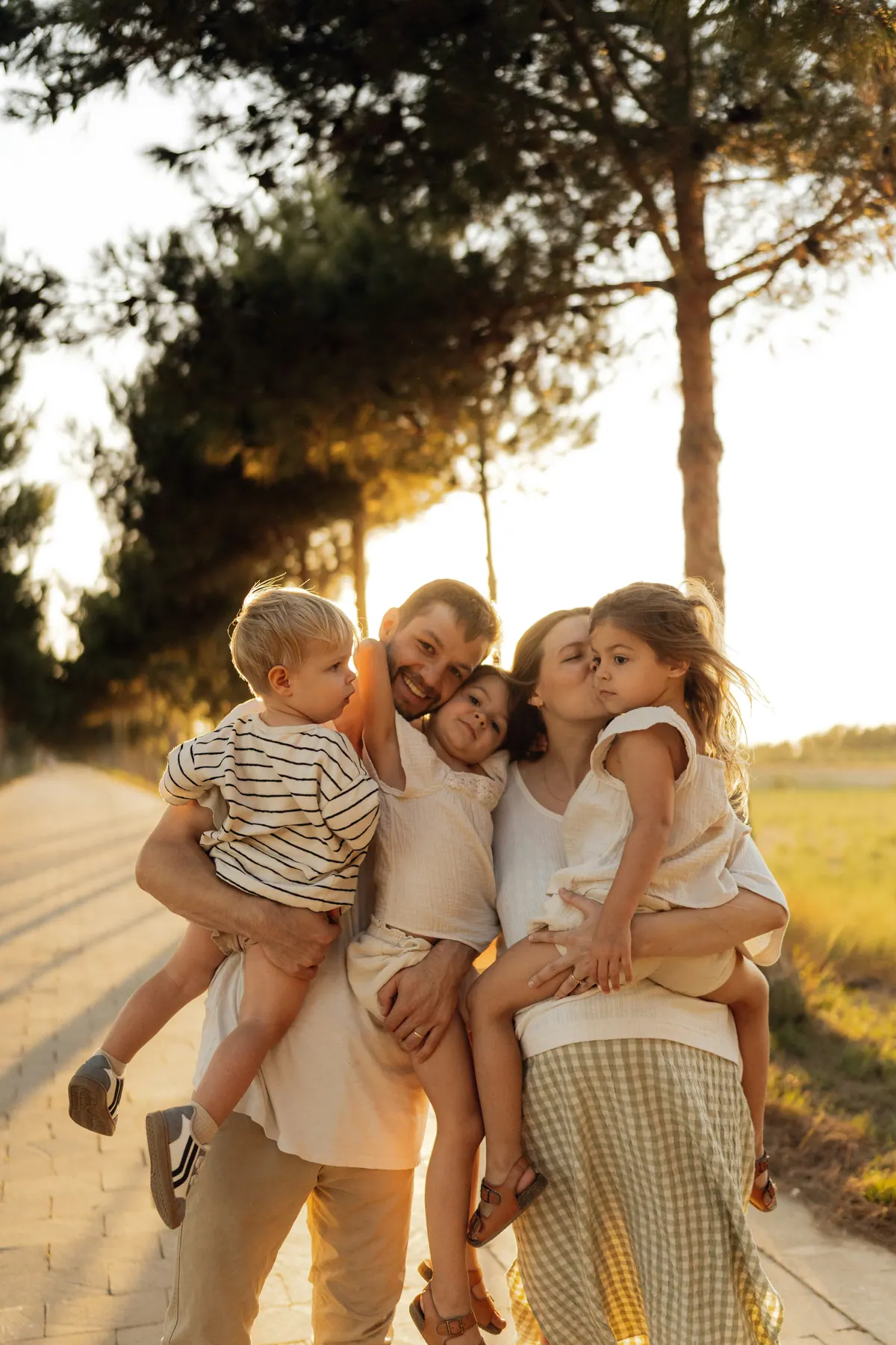 Two parents each carrying a child along a tree lined path in evening light