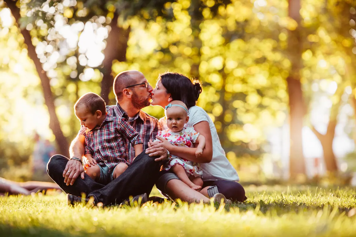 Two parents and two young children sitting together on sunlit grass under trees