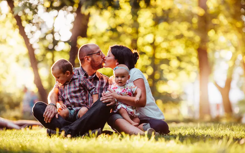 Two parents and two young children sitting together on sunlit grass under trees