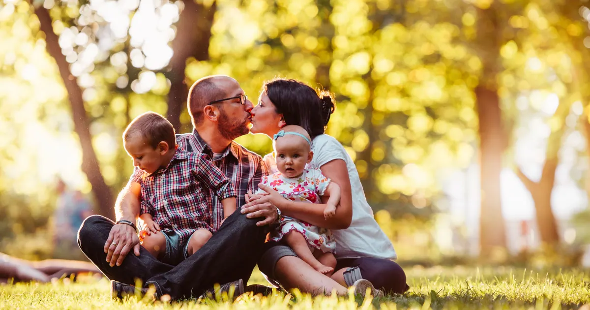 Two parents and two young children sitting together on sunlit grass under trees