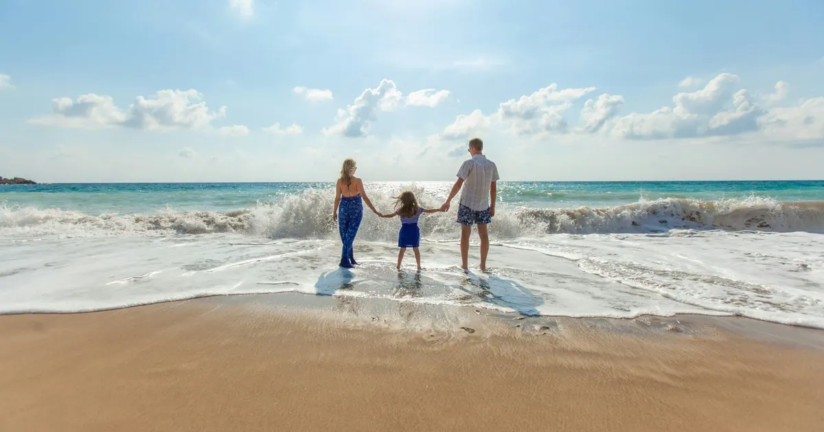 Two parents and a child holding hands in shallow surf on a wide sandy beach