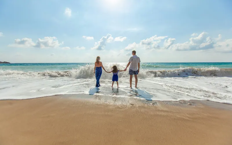 Two parents and a child holding hands in shallow surf on a wide sandy beach