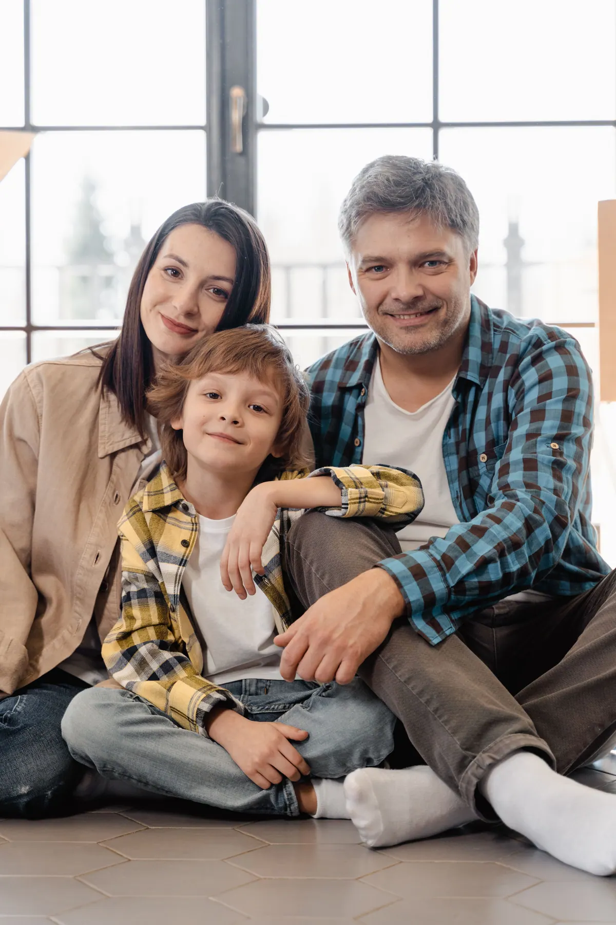 A mother, father and young son sitting together on the floor beside a tall window at home
