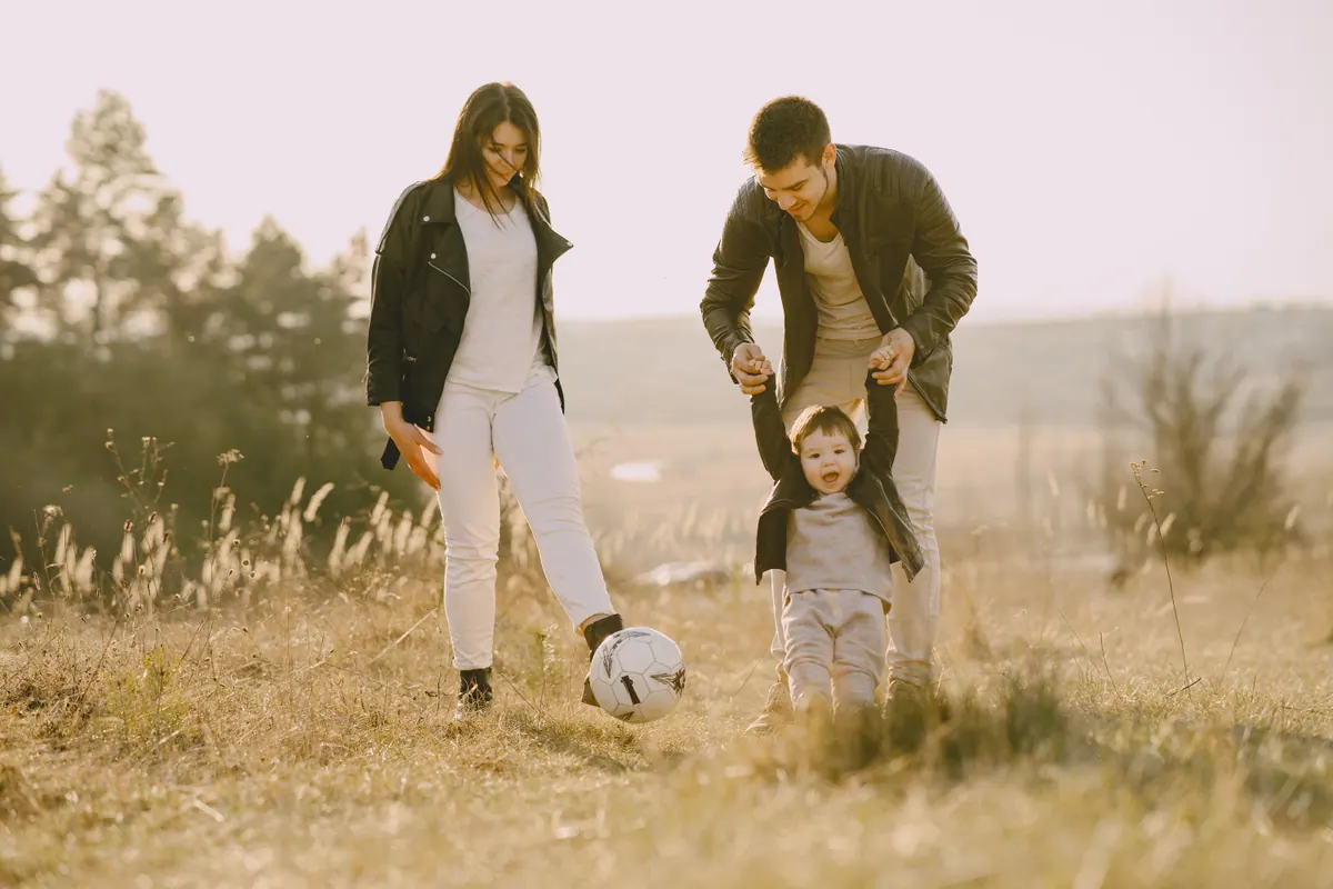 Two parents and a small child playing with a football in a dry grass field