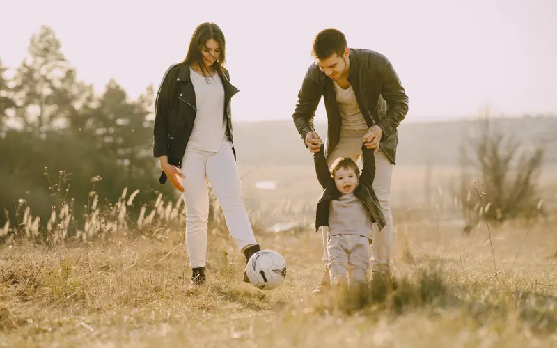Two parents and a small child playing with a football in a dry grass field