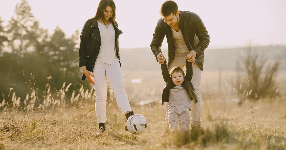Two parents and a small child playing with a football in a dry grass field