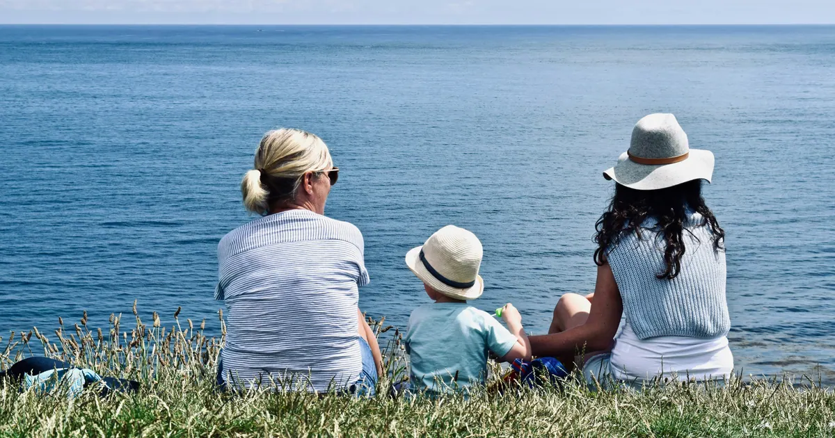 Two adults and a small child sitting on grass, looking out over open water