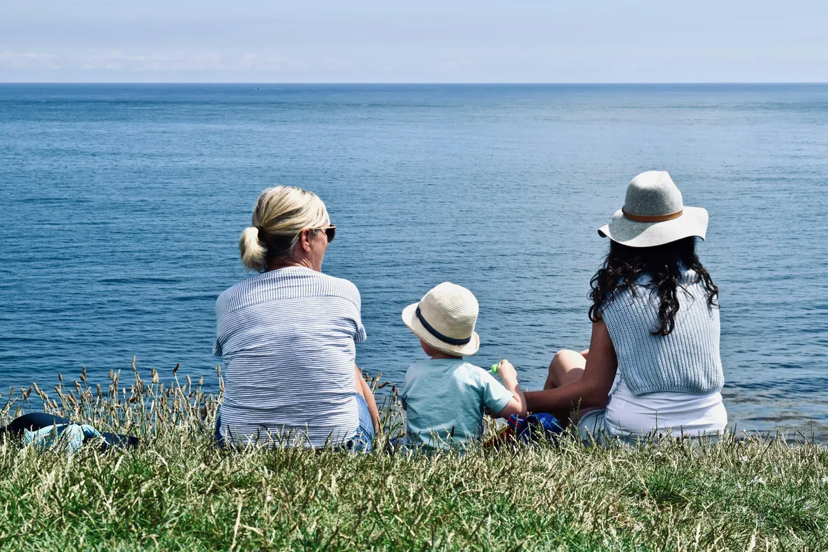 Two adults and a small child sitting on grass, looking out over open water