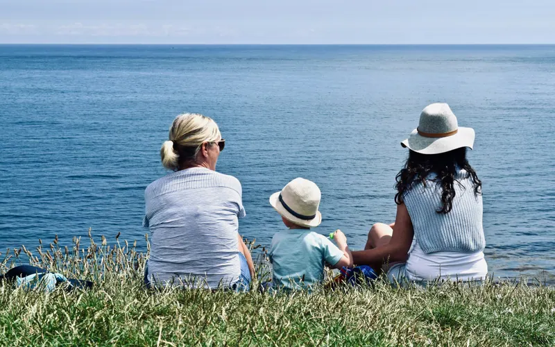 Two adults and a small child sitting on grass, looking out over open water