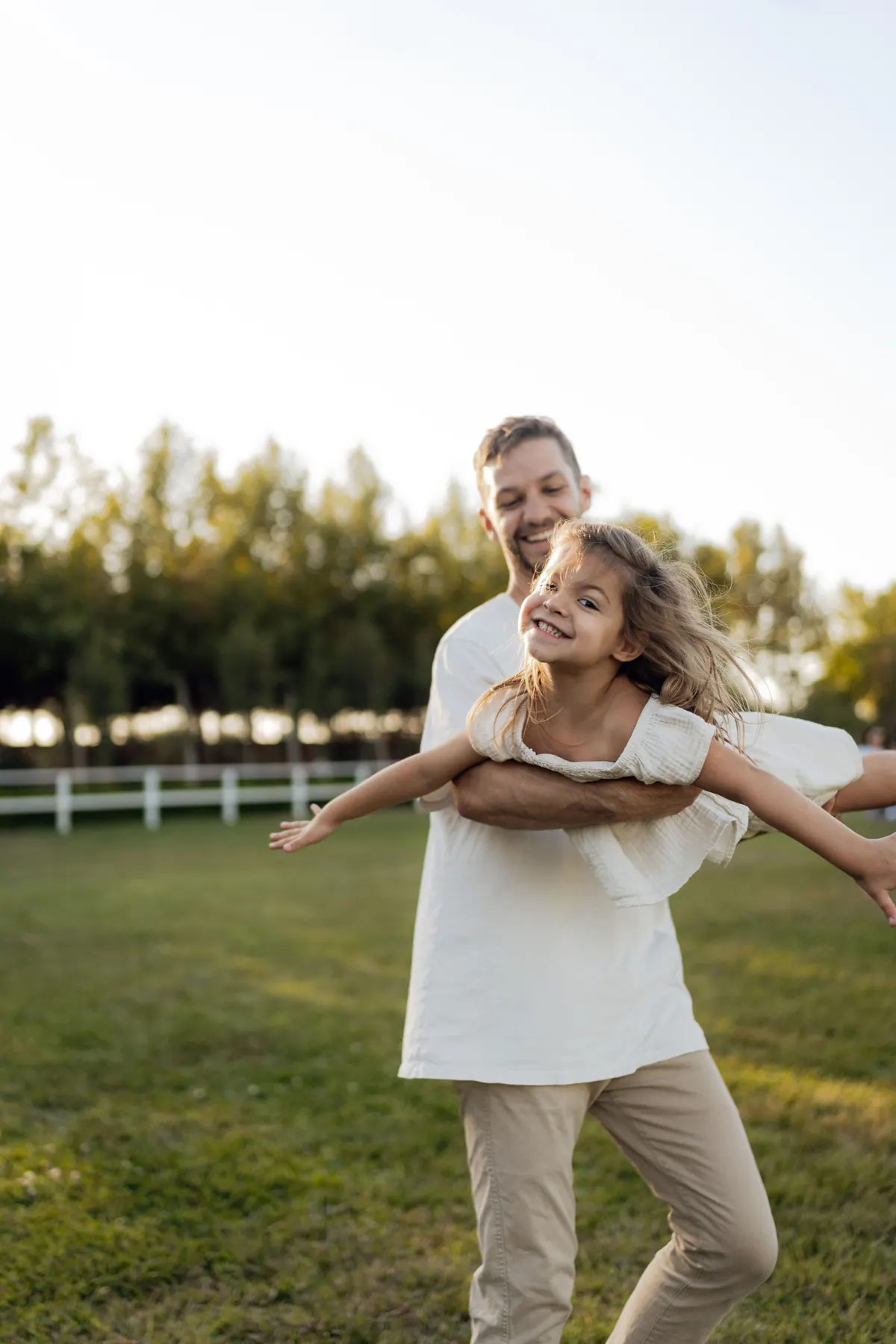 A father carrying his daughter across a field with her arms stretched out