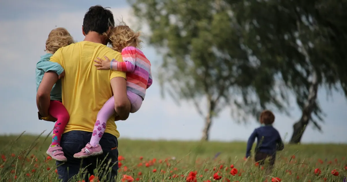A father carrying two young children through a field of poppies, a third child running behind