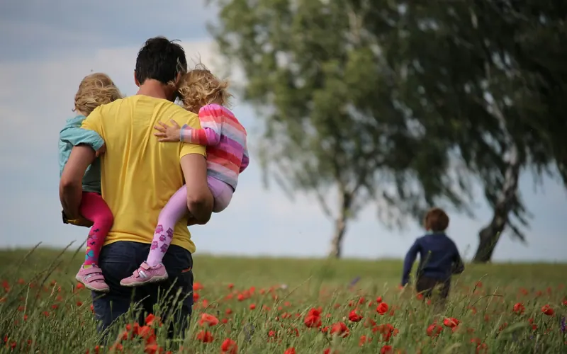 A father carrying two young children through a field of poppies, a third child running behind