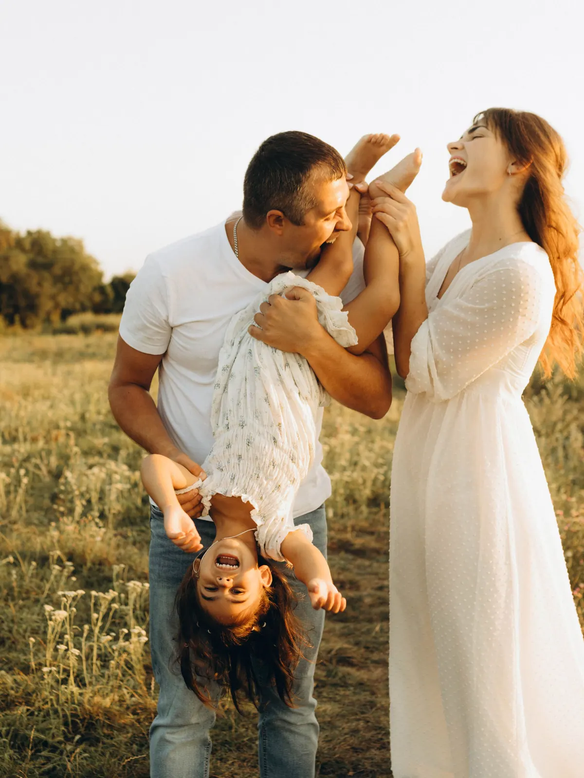 A father holding a laughing child upside down while the mother laughs beside them in a meadow