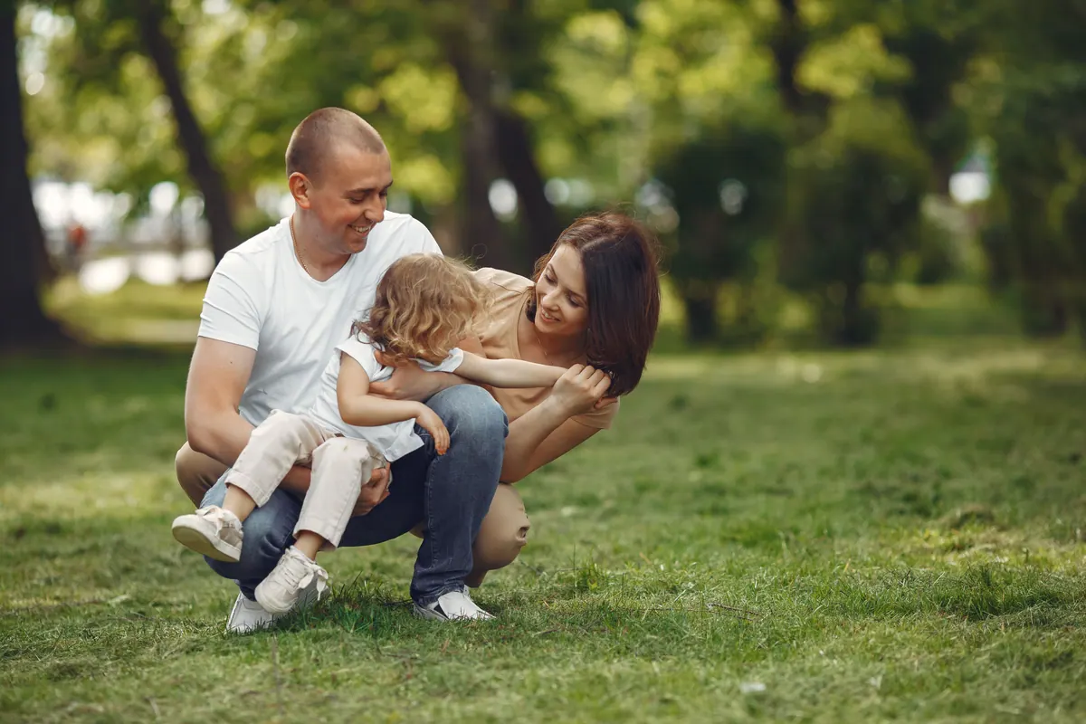 A father kneeling on the grass holding his son while the mother leans in, in a green park