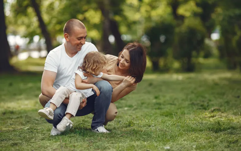 A father kneeling on the grass holding his son while the mother leans in, in a green park