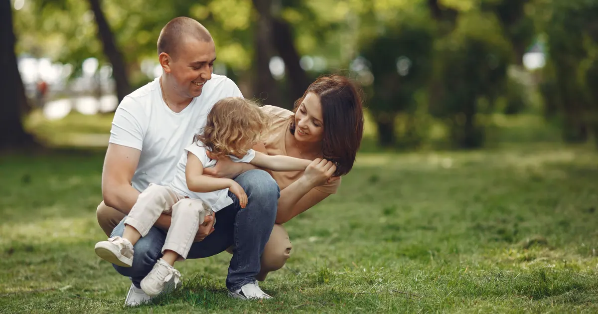 A father kneeling on the grass holding his son while the mother leans in, in a green park