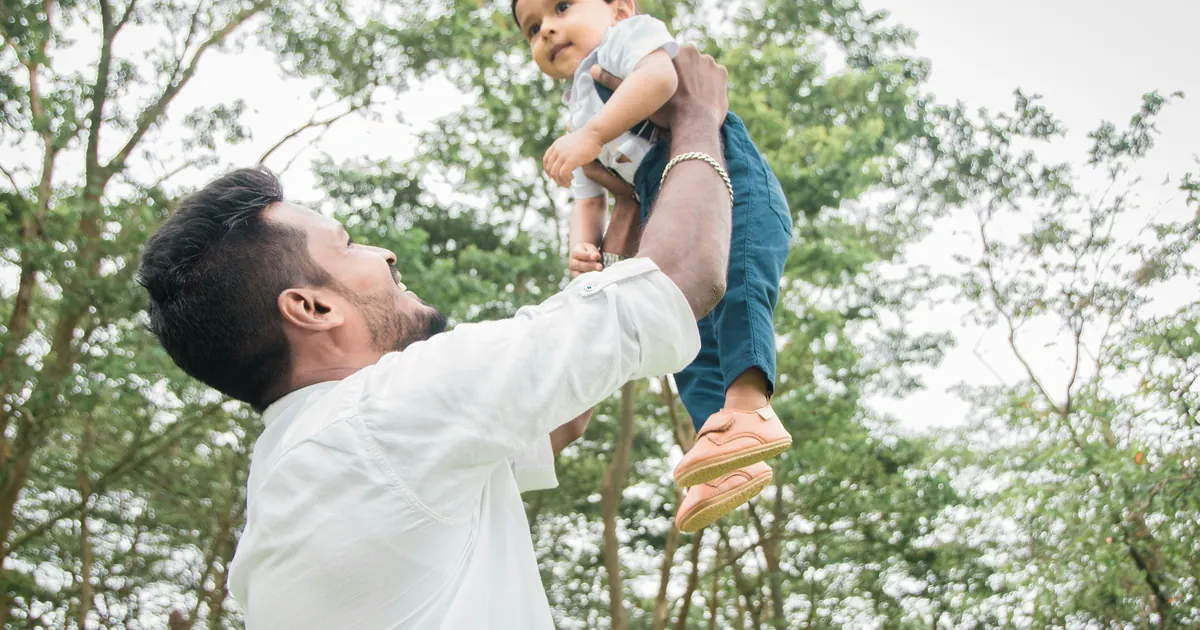 A father lifting a laughing toddler above his head beneath green trees