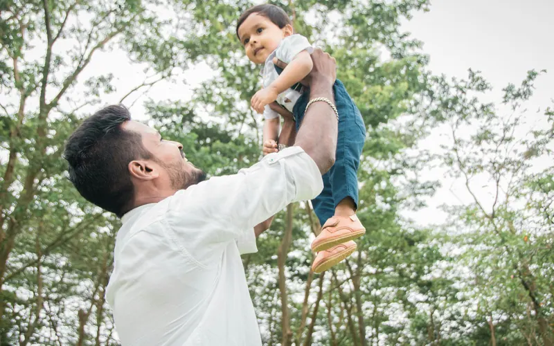 A father lifting a laughing toddler above his head beneath green trees