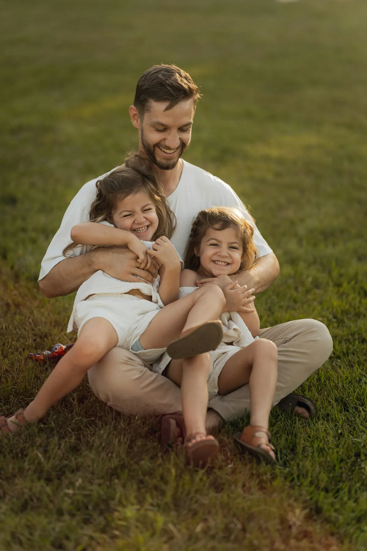 A father sitting on the grass with two young daughters leaning into him