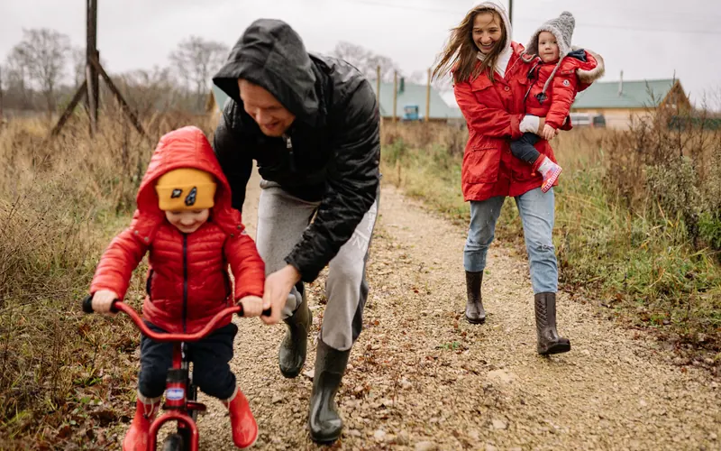 A father steadying a small child on a red bicycle on a rural gravel track, the mother carrying another child behind