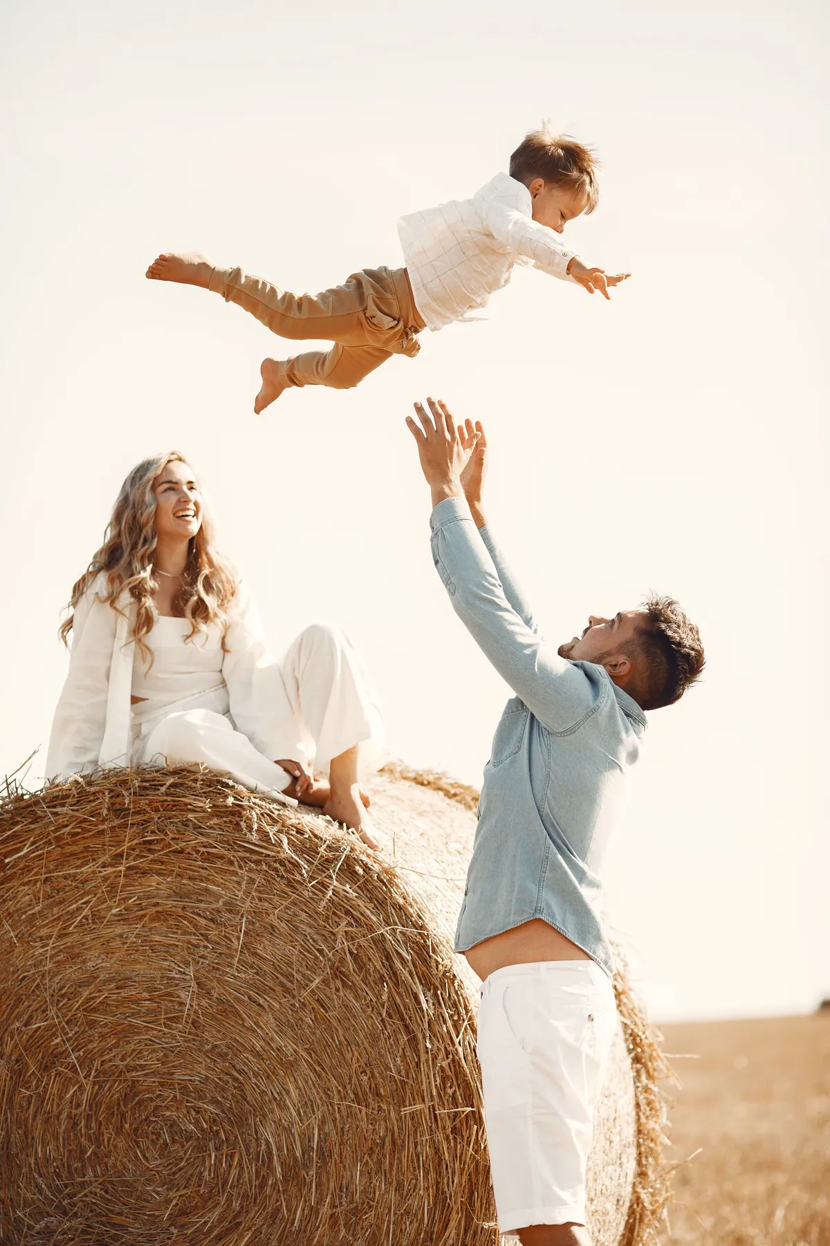 A father tossing a small child into the air above a hay field while the mother watches