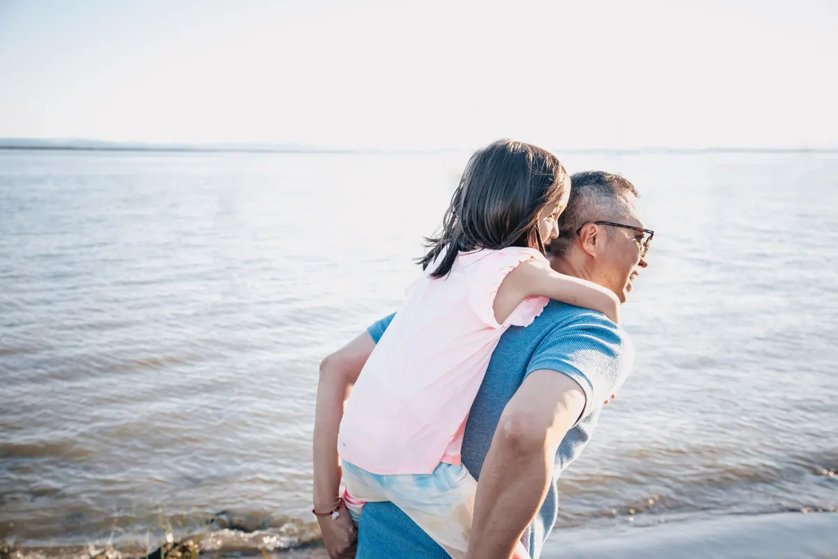An older man carrying a laughing child on his back beside open water