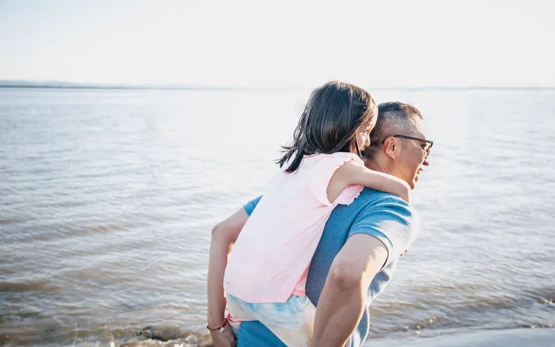 An older man carrying a laughing child on his back beside open water