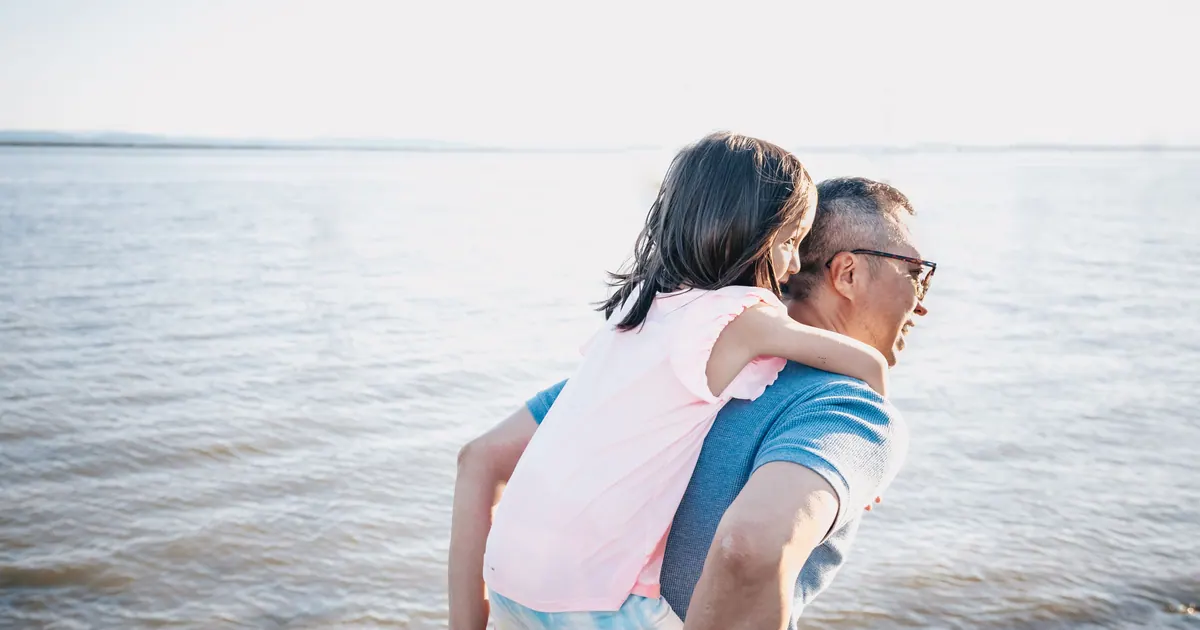 An older man carrying a laughing child on his back beside open water
