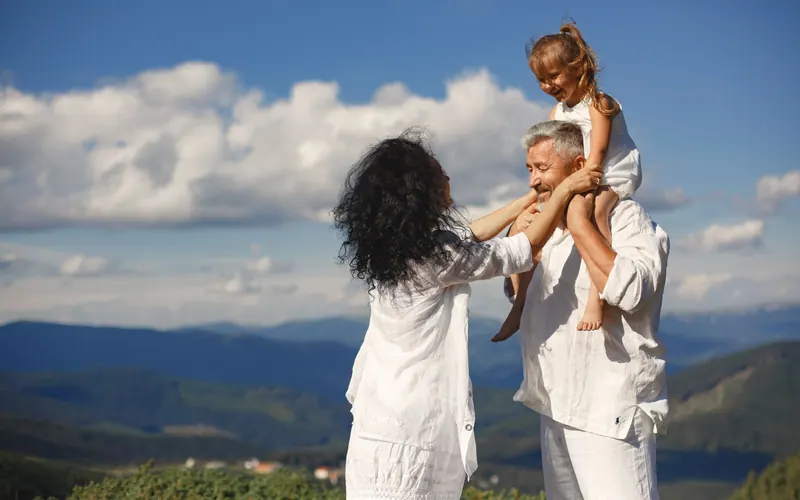 An older couple with a young girl on the man's shoulders, mountains and cloud behind them