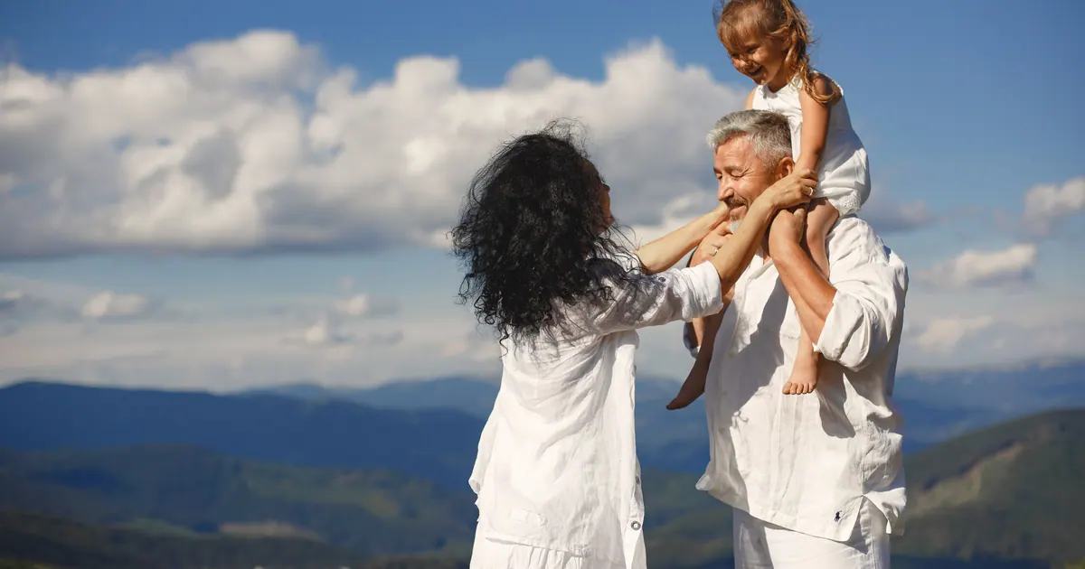An older couple with a young girl on the man's shoulders, mountains and cloud behind them