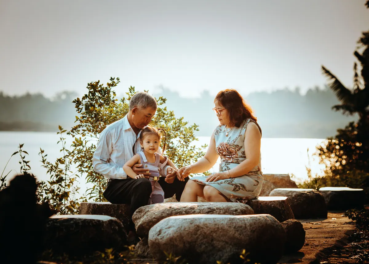An older couple sitting on rocks beside a lake with a toddler standing between them