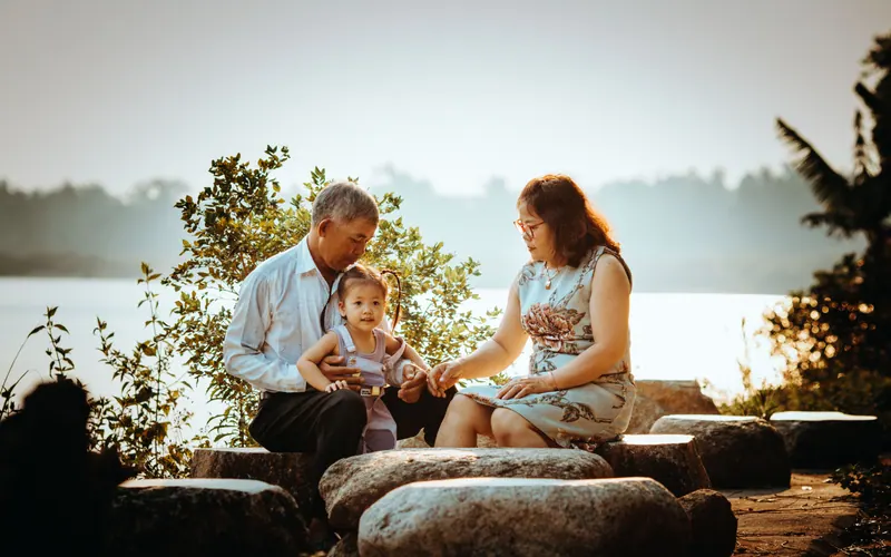 An older couple sitting on rocks beside a lake with a toddler standing between them
