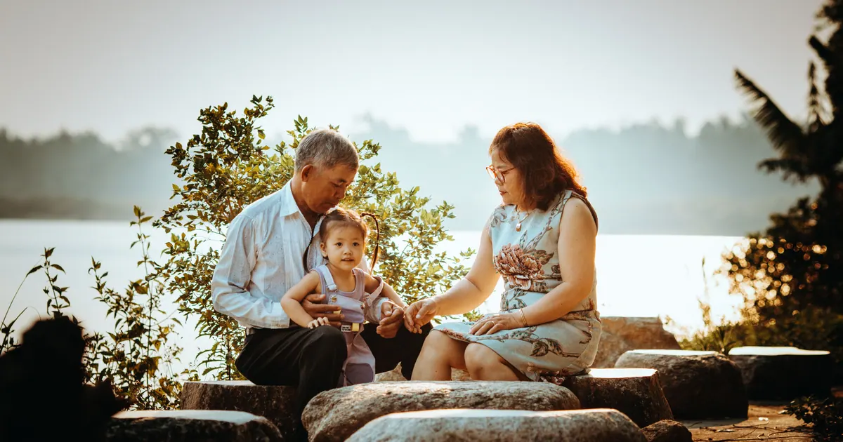 An older couple sitting on rocks beside a lake with a toddler standing between them