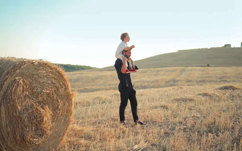 A man carrying a small child on his shoulders in a field of round hay bales